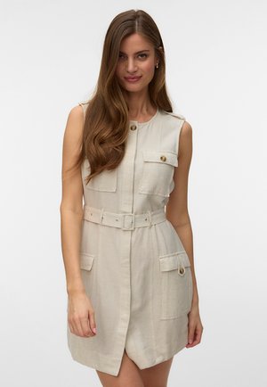 Vrouw met lang bruin haar, gekleed in een mouwloze beige jurk met riem, voorzakken en knopen, staand tegen een effen achtergrond.