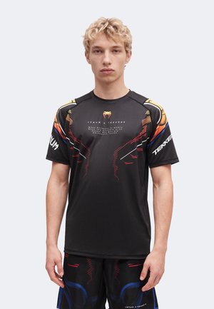 Młody mężczyzna w czarnej odzieży sportowej Venum x Tekken 8 z pomarańczowymi, czerwonymi, niebieskimi i białymi akcentami graficznymi, stojący na jednolitym tle.