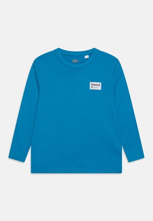 Langärmliges Hemd aus leuchtend blauem Baumwollstoff, mit Rundhalsausschnitt und kleinem weißen Timberland-Logo-Patch auf der linken Brust. Samtige Textur.