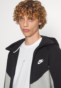 Nike zip hoodie i sort og grå med en blød tekstur. Har et hvidt logo på brystet og en kontrasterende hvid t-shirt underneath.