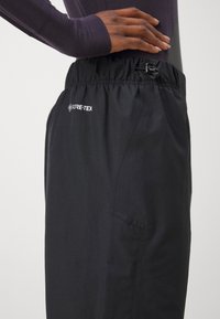 Crne GORE-TEX hlače s elastičnim pojasom, koje imaju zatvaranje na vezanje, glatku teksturu i bočnu dzep.