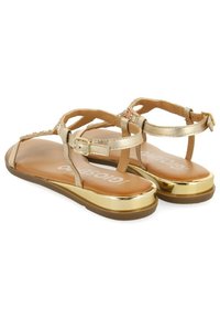 Sandales plates dorées avec des brides à la cheville, une semelle intérieure en tan rembourrée et un accent doré métallique sur la semelle. Décoration en perles sur les brides.