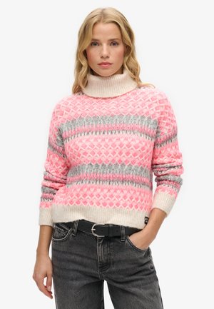 Lyshåret kvinde iført en lyserød og grå mønstret rullekravtrøje med sorte jeans, poserer med den ene hånd i lommen.