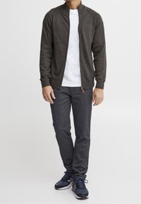 Man som bär en mörkgrå zip-up cardigan, vit t-shirt, mörkgrå byxor och marinblå sneakers, stående mot en vit bakgrund.