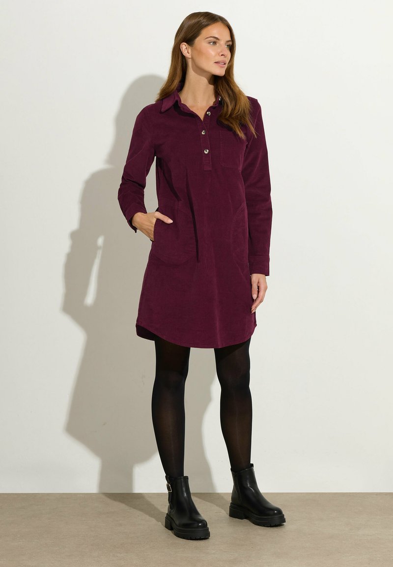 Burgunderfarbendes Cordkleid mit einem Knopfkragen, langen Ärmeln und seitlichen Taschen. Kombiniert mit schwarzen Knöchelschuhen. Glatte Textur, figurbetonte Form.