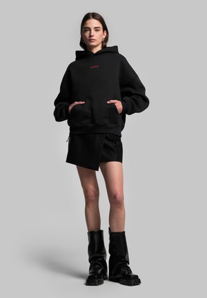 Black oversized hoodie with front pouch pocket, featuring red text. Paired with a black asymmetrical skirt and tall black boots.