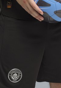 Hånd, der holder en blå og sort Nike fodbold over sorte Manchester City shorts med klubmærke og Puma branding.