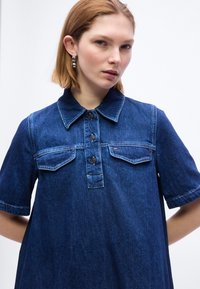 Μια νεαρή γυναίκα φοράει μια κοντομάνικη σκούρα μπλε τζιν πουκάμισο με κουμπιά μπροστά και τσέπες με καπάκια, στέκεται μπροστά σε ένα απλό λευκό φόντο.