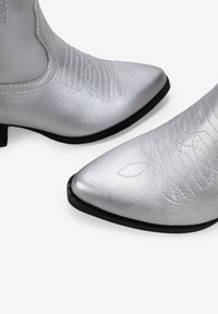 Botas de vaquero plateadas con puntas afiladas, textura metálica suave, bordados intrincados y suela negra. Sin hardware visible.