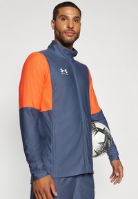 Marineblaues und orangefarbenes sportliches Zip-Jackett mit hohem Kragen und langen Ärmeln. Mit Logo unter dem Arm und strukturiertem Stoff. Hält einen Fußball.