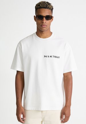 Junger Mann mit kurzen lockigen Haaren, der dunkle Sonnenbrillen und ein weißes T-Shirt mit "YOU & ME TONIGHT" auf der Brust trägt.