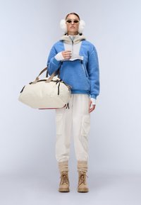 Blue fleece jacket, off-white cargo pants, beige boots; carrying a cream duffel bag; includes round sunglasses and white ear muffs.