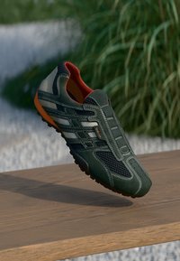 Zielone zamszowe sneakersy z siateczkowymi panelami, szarymi akcentami i czerwonym wnętrzem. Posiadają teksturowaną gumową podeszwę i przeszycia.