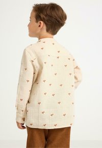 Beige Langarmhemd mit verstreuten Fuchs-Motiven, ausgestattet mit einem Knopfmanschette und einer lässigen Passform. Kombiniert mit braunen Cordhosen.