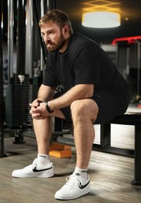 Zwart shirt met korte mouwen, zwarte shorts, witte Nike Air Force 1-schoenen met zwart swoosh, zittend op een gymbank in een binnenomgeving.