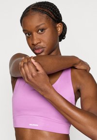 Ανοιχτό μωβ αθλητικό σουτιέν με λείο υφή, με εφαρμοστή σχεδίαση και λογότυπο στο κάτω μέρος. Το μοντέλο κρατάει το χέρι της, παρουσιάζοντας τη εφαρμογή.