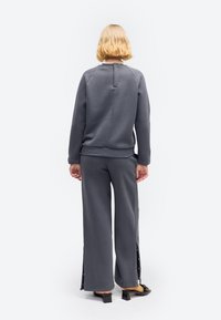 Person med kort blondt hår iført en mørkegrå sweatshirt og vidde bukser med slidser i siderne, stående med ryggen mod kameraet.