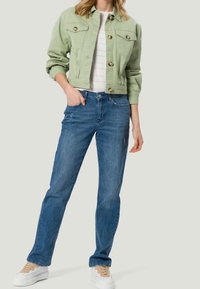 Lysgrøn cropped denimjakke med to forlommer og messingknapper, parret med blå straight-leg jeans og hvide sneakers.