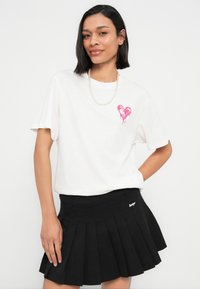 Άσπρο βαμβακερό t-shirt με ροζ καρδιά στο αριστερό στήθος, συνδυασμένο με μαύρη πλισέ φούστα. Απλός σχεδιασμός, άνετη εφαρμογή.