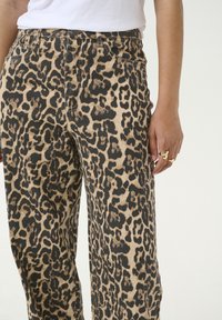 Pantalons à imprimé léopard en beige et noir, avec une taille haute, des poches latérales et un design à jambe droite. Des bagues dorées sont visibles sur la main.