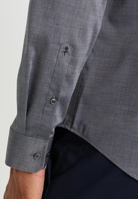 Grå, texturerad skjorta med långa ärmar, knäppningar vid ärmsluten och en broderad rosdetalj på ärmslutet. Har en figurnära design.