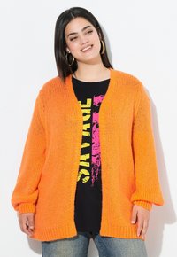 Cardigan orange en tricot avec des manches longues, design ouvert à l'avant et ourlet côtelé. Porté sur une chemise noire avec un imprimé graphique coloré.