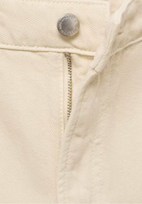 Gros plan sur un tissu en denim beige montrant une fermeture éclair argentée partiellement ouverte et un bouton avec un texte gravé.
