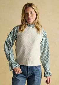 Blonde Frau, die ein cremripiertes Strickoberteil über einer grün-weiß gestreiften Bluse mit Rüscheneinsätzen und blauen Jeans trägt.