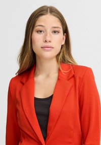 Blazer ajusté rouge avec des revers crantés, des épaules structurées et une texture lisse, superposé sur un débardeur noir en maille côtelée.