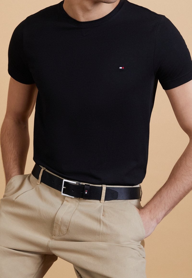 Schwarzes T-Shirt mit kurzen Ärmeln und Rundhalsausschnitt, mit einem kleinen rot-, weiß- und blauem Logo auf der linken Brust. Getragen mit beigen Hosen und einem schwarzen Gürtel.