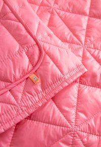 Roze doorgestikte stof met diamantstiksels en een omgevouwen hoekje waarop een oranje label met een klein grijs hondenlogo te zien is.