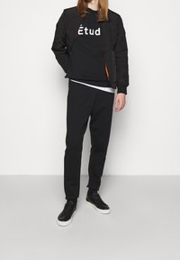 Chaqueta bomber acolchada negra, sudadera negra con el texto "Étud", pantalones de chándal negros y zapatillas negras con suelas blancas, sobre un fondo liso.