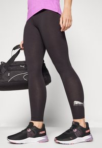 Svarta leggings med en glänsande silverfärgad Puma-logotyp på sidan, i kombination med svarta och rosa träningsskor, hållna med en svart gymväska.