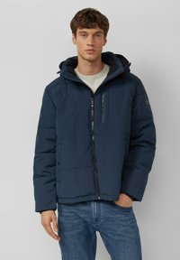 Granatowa kurtka puchowa z zamkiem z przodu, regulowanym kapturem i pikowaną teksturą. Posiada kieszeń na piersi i elastyczne mankiety. Połączona z dżinsami.