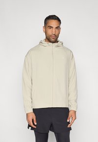 Könnyű, bézs színű cipzáras kabát kapucnival. Rugalmas mandzsetták és egy minimalista logó jellemzi. Sötét rövidnadrág fölött viselve, sima anyagtextúrával.