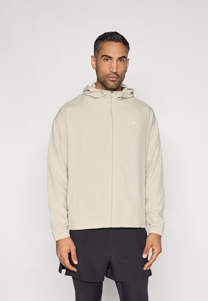 Lekka, beżowa kurtka na zamek z kapturem. Posiada elastyczne mankiety i minimalistyczne logo. Nosi się ją na ciemnych szortach, gładka tekstura tkaniny.