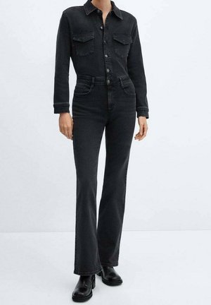 Personne portant une combinaison en jean noire à manches longues, poches poitrine, boutonnée sur le devant, jambes évasées et bottines noires, sur un fond uni.