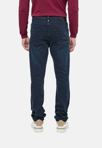 Homme portant un jean bleu foncé, des baskets blanches et une chemise à manches longues bordeaux, se tenant de dos contre un fond uni.