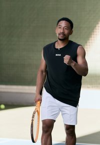 Juodas be rankovių sportinis marškinėlis su "BOSS" logotipu, balti šortai, laikantis oranžinį teniso raketę, ant aikštelės su žaliu tinklu fone.