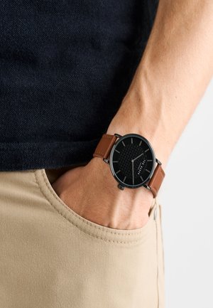 Pruun nahast rihmaga kell, millel on must ümmargune kellanägu koos tekstureeritud mustriga, hõbedased tunni markerid ja must metallkorpus.