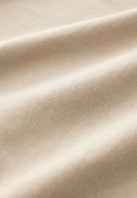 Beige katoenen stof met een gladde textuur en een subtiele glans, met plooien en zachte golven die een zacht, vloeiend uiterlijk creëren.