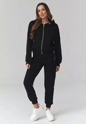 Kobieta z długimi włosami, ubrana w czarną bluzę na zamek i pasujące spodnie dresowe, stojąca na tle jednolitego szarego tła w białych tenisówkach.