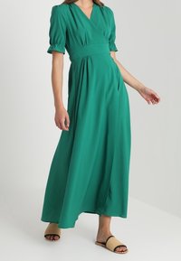 Vestido maxi verde con un diseño envolvente, mangas cortas abullonadas y cintura ceñida. Hecho de una tela suave, presenta una abertura lateral y una silueta fluida.