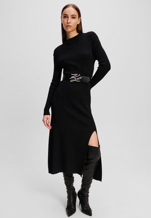 Črna rebrasta pletena obleka z dolgimi rokavi, sprednjim razporkom in širokim črnim pasom z okrasnimi biseri. Manekenka nosi črne škornje do stegen.