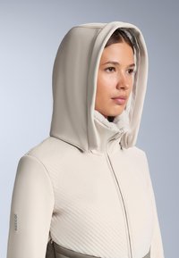 Bézs kapucnis dzseki steppelt mintázattal, sima ujjakkal és Recco® márkajelzéssel. Puha bélés a gallér és a kapucni körül a kényelem érdekében.