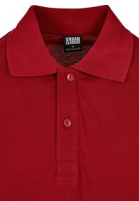 Rød polo skjorte laget av teksturert stoff, med spiss krage og to røde knapper. Merket viser "URBAN CLASSICS," størrelse M, laget i Bangladesh.