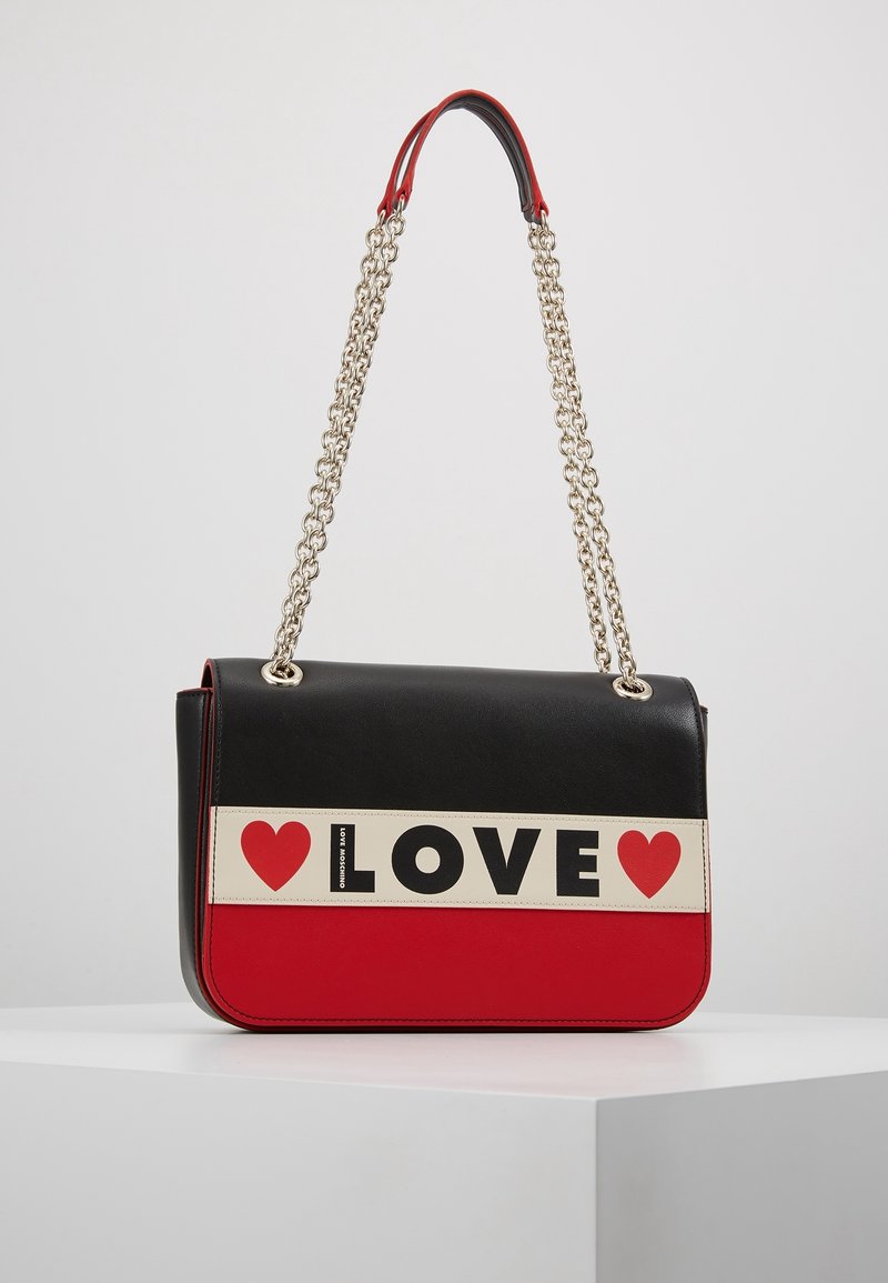 Černá a červená kabelka s řetízkem, která má krémový pruh s nápisem "LOVE" tučným černým písmem a ikonami červeného srdce. Hladký povrch.