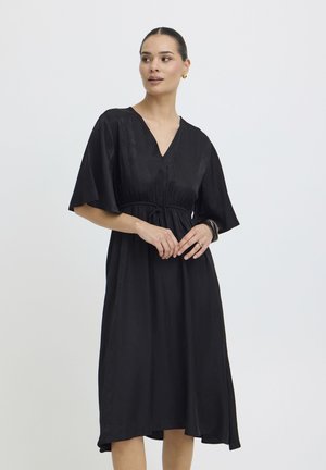 Naine, kellel on juuksed tagant seotud, kannab musta V-kaelusega midi-kleit lahtiste varrukatega, vöökoha peal kitsendatud, kuldsete kõrvaklappidega ja musta käevõruga, vaadates küljele.