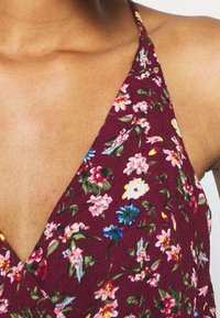 Burgundské květinové sluneční šaty s výstřihem do V, zdobené malými růžovými, žlutými a modrými květy se zelenými listy, vyrobené z lehké tkaniny.