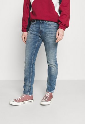 Personne portant un jean slim bleu délavé, un sweat-shirt rouge et noir, et des baskets montantes en toile rose, debout sur un sol clair uni.